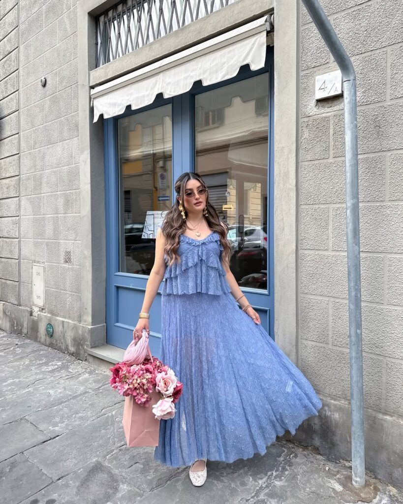 Mulher jovem de pele clara e cabelo castanho. Ela usa blusa e saia azul, sapatilha e segura uma bolsa com flores