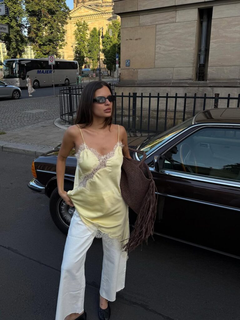 Mulher jovem, magra e de pele clara com cabelo castanho. Ela usa blusa acetinada com renda, calça jeans branca e sapatilha preta