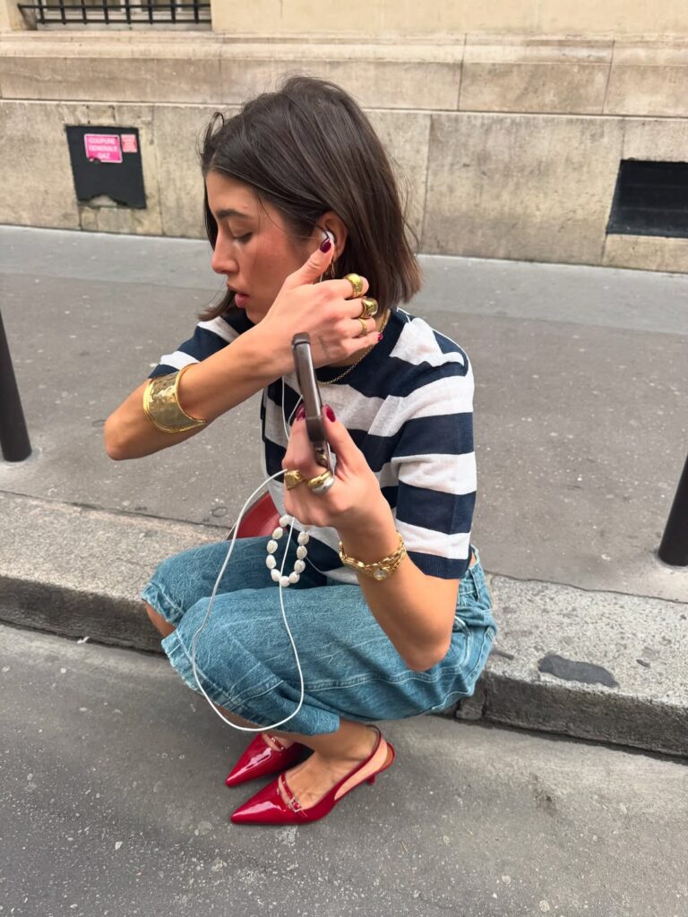 Mulher jovem, magra e de pele clara. Ela tem cabelo curto castanho e usa blusa listrada, bermuda e scarpin slingback vermelho