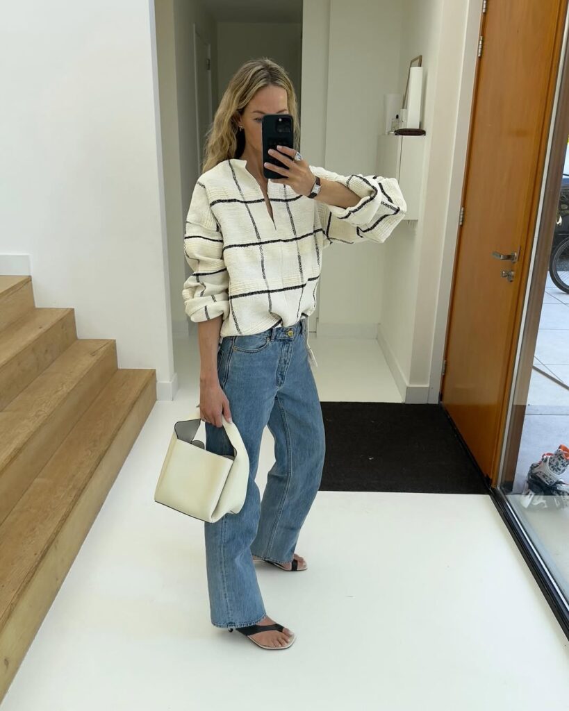 Mulher jovem, magra e de pele clara. Ela tem cabelos loiros e está tirando uma foto na frente do espelho. Ela veste camisa, calça jeans e sandália.