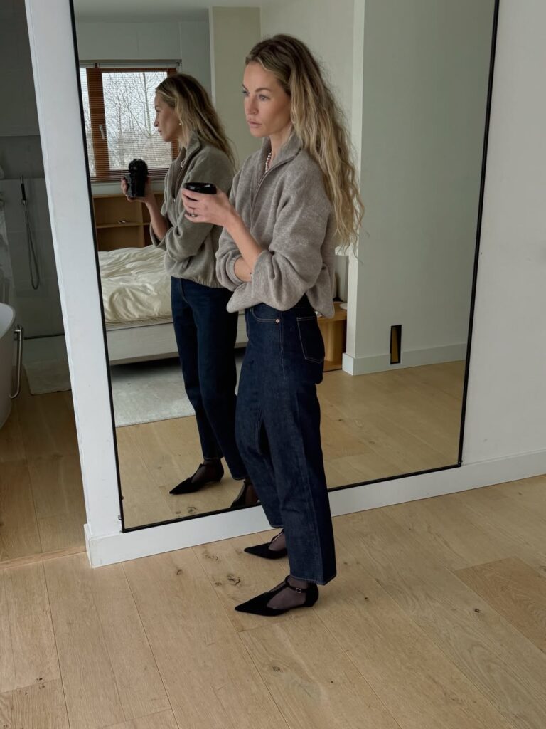 Mulher jovem, magra e de pele clara. Ela usa suéter, calça jeans escura e sapatos pretos