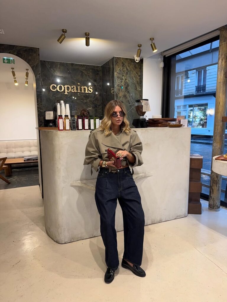 Mulher jovem e magra com pele clara e cabelos loiros. Ela usa casaco, calça jeans e mocassim