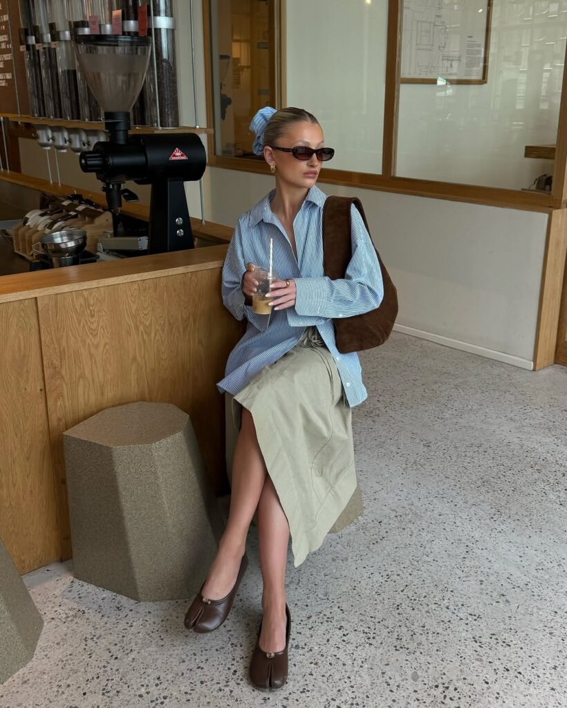 Mulher jovem e magra de pele clara. Ela usa camisa de manga longa azul, saia midi cinza e sapatilha marrom