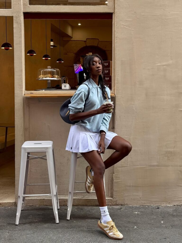 Mulher jovem, magra, alta e de pele negra. Ela usa suéter, minissaia, tênis com meia e segura uma bolsa