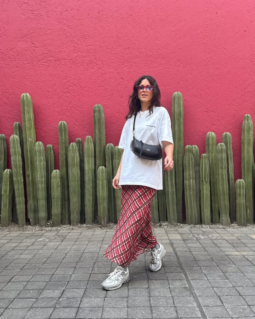 Mulher jovem com look com camiseta branca oversized, saia midi xadrez, tênis esportivo e bolsa transversal pequena