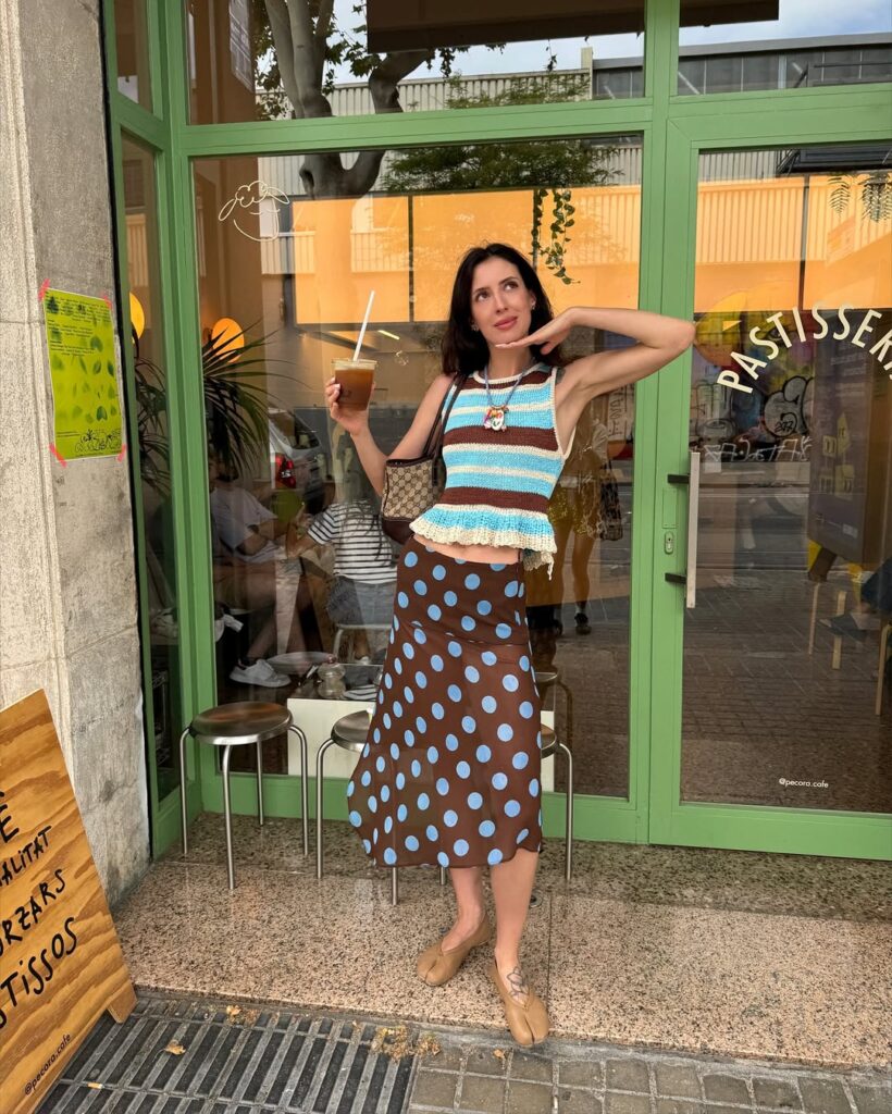 Mulher jovem e magra de pele clara com cabelos castanhos. Ela usa blusa com listras, saia midi de poá, sapato tabi e bolsa de ombro