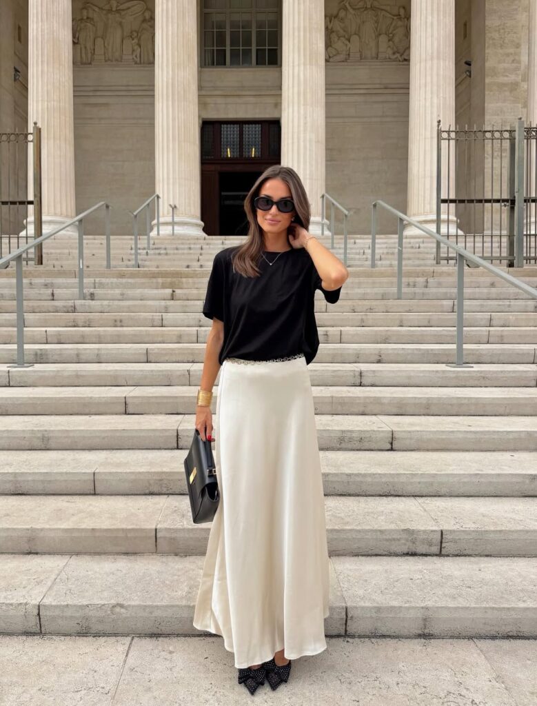 Mulher jovem, magra e de pele clara com cabelo castanho médio. Ela usa camiseta preta, saia de cetim feminina lonfa e sapato de bico fino com laços