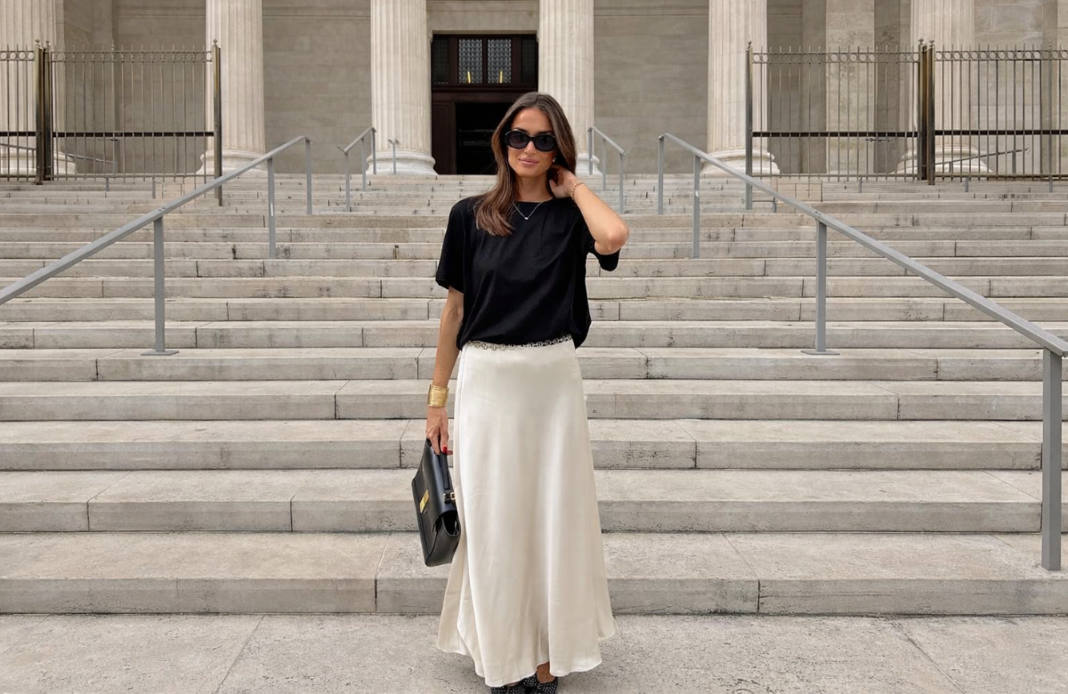 Mulher jovem, magra e de pele clara com cabelo castanho médio. Ela usa camiseta preta, saia de cetim feminina lonfa e sapato de bico fino com laços
