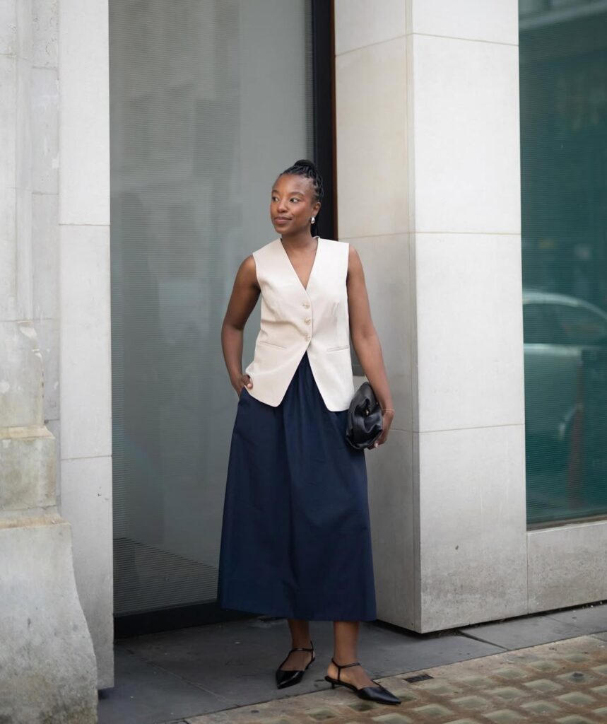 Mulher jovem de pele negra. Ela usa colete de alfaiataria, saia midi e salto alto.