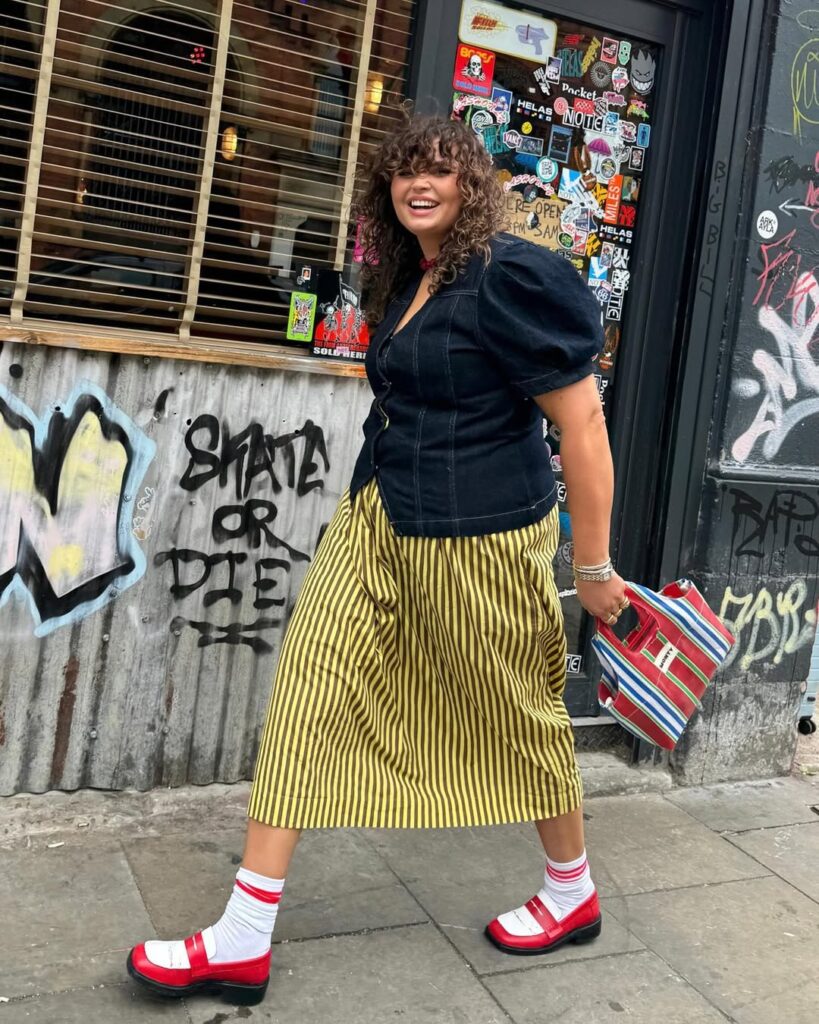 Mulher jovem de pele clara com cabelo curto e cacheado. Ela usa blusa jeans com manga bufante, saia midi com listras, mocassim com meia e segura uma bolsa com listras coloridas.